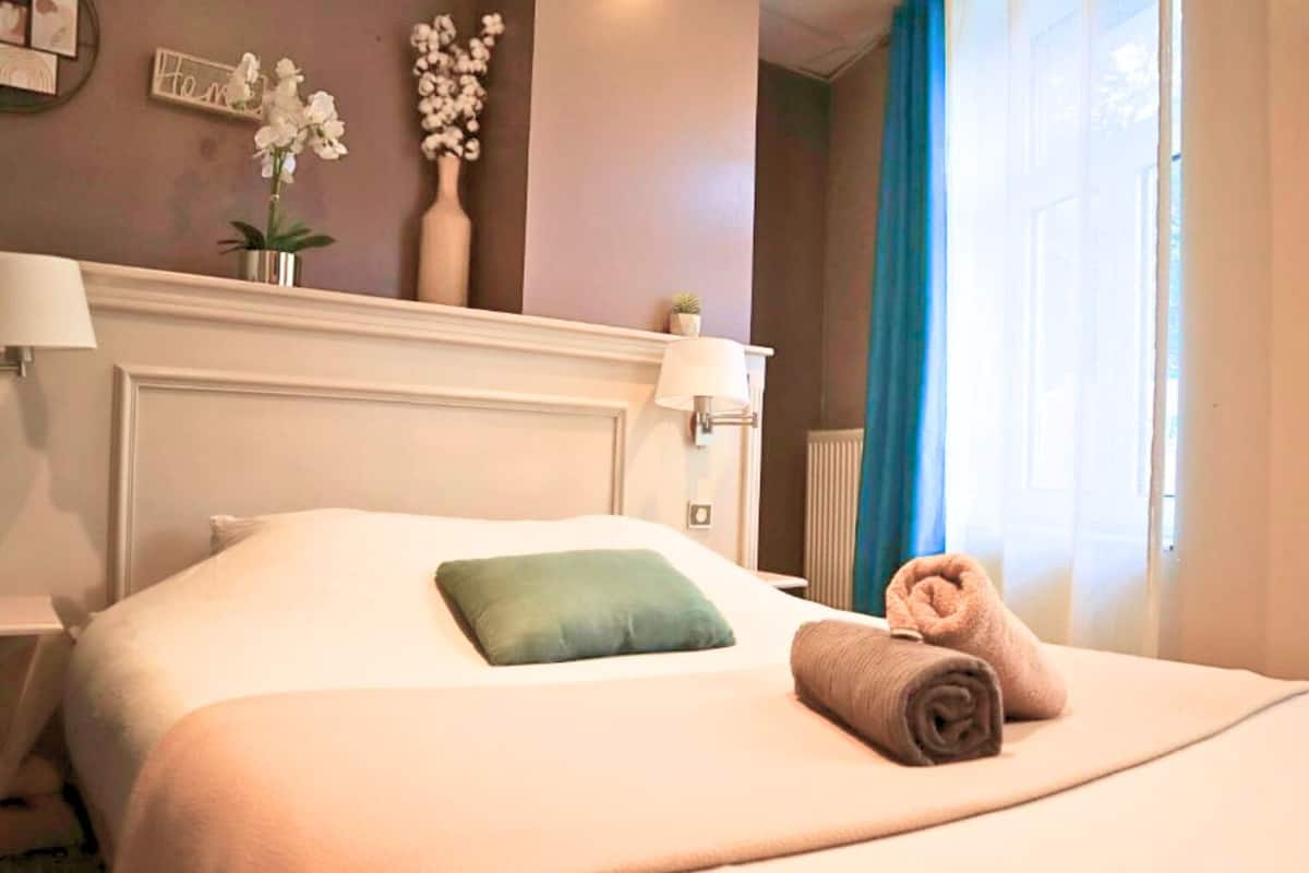 Una cama bien hecha con una almohada verde y una toalla enrollada, una mesita de noche con una lámpara, un jarrón con flores y una pared con un cartel que dice 'Home'.