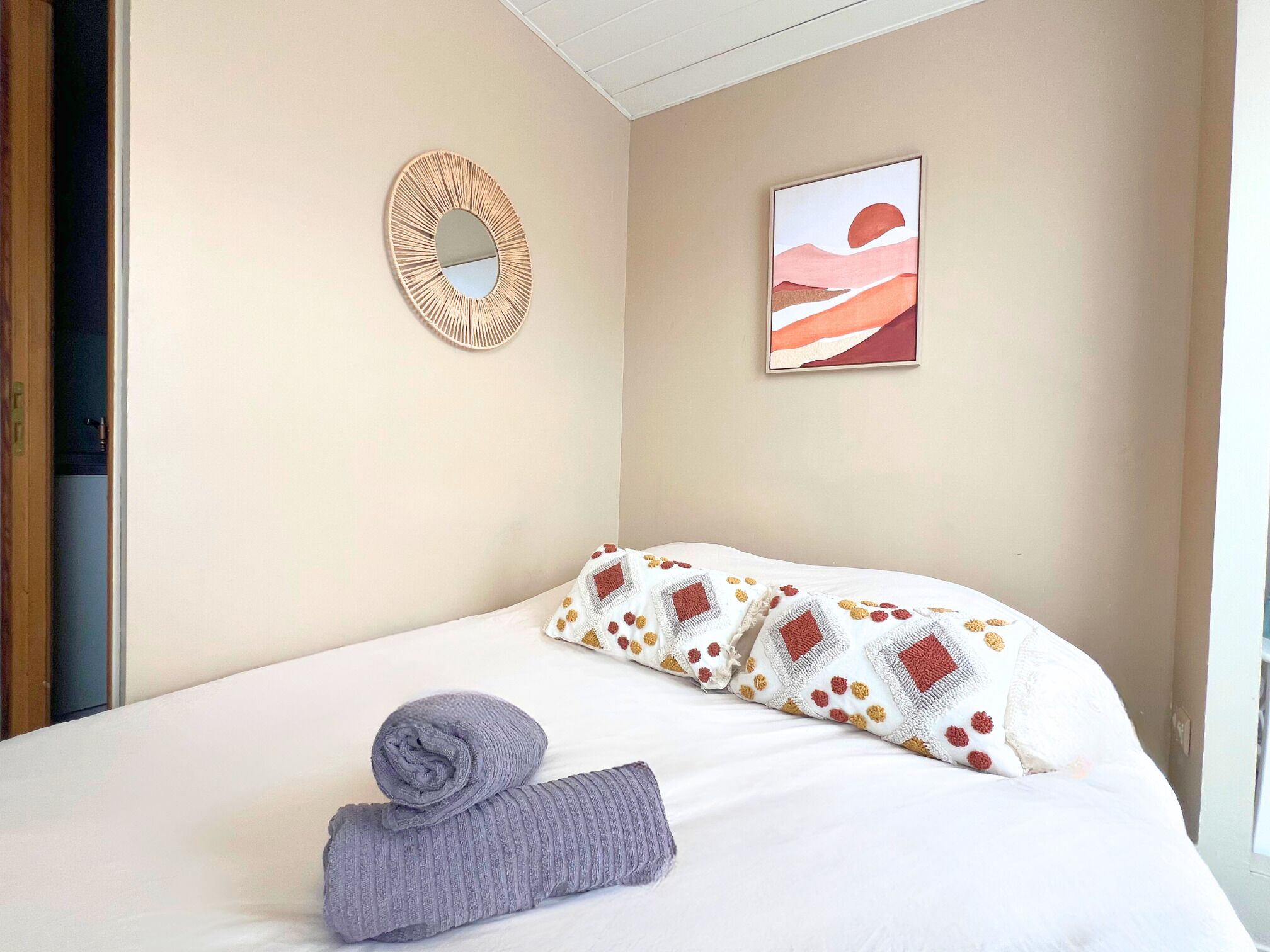 Una cama bien hecha con un cojín estampado y una toalla enrollada, un espejo montado en la pared y un cuadro abstracto enmarcado.