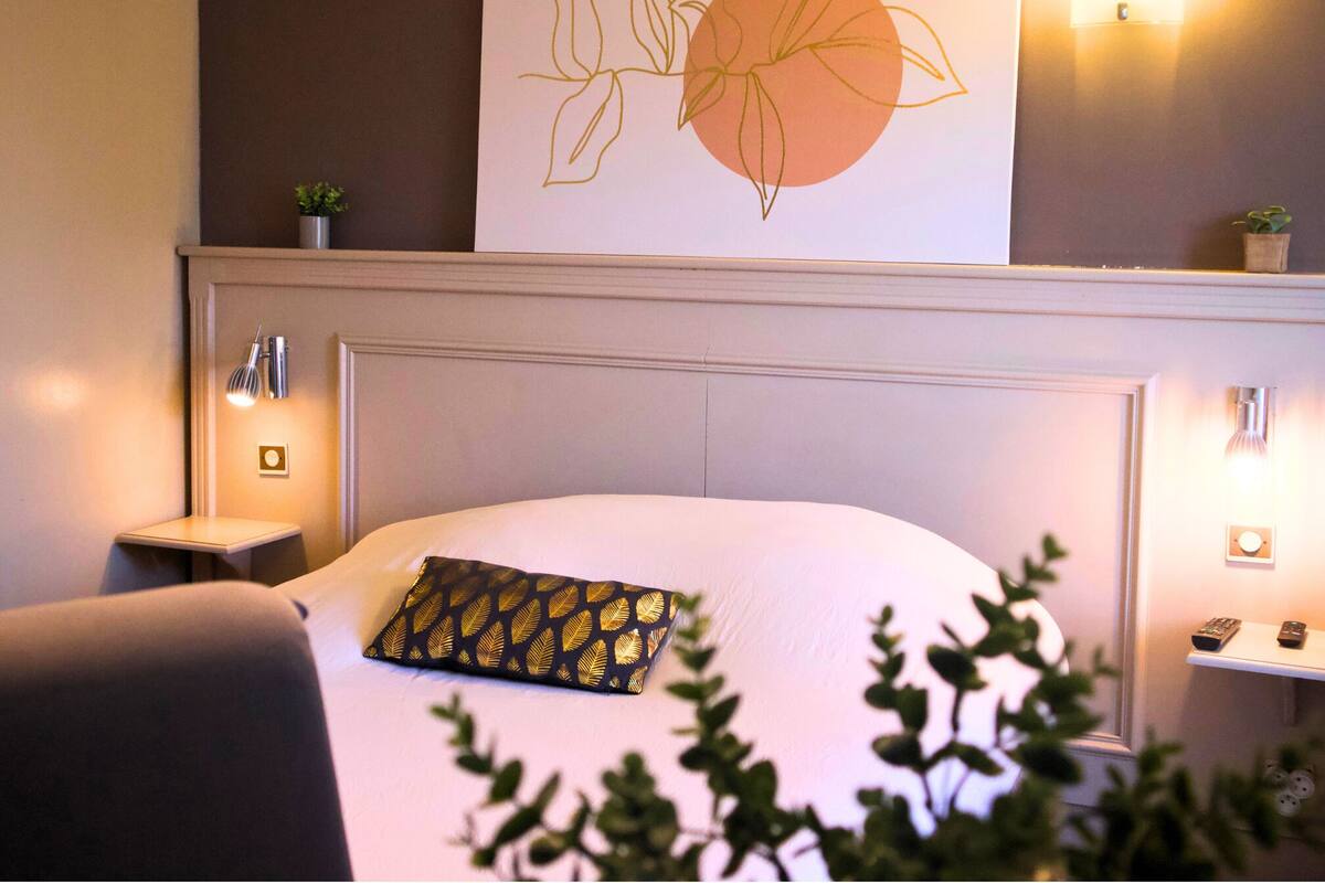 Una habitación de hotel con una cama, cabecero, luces fijadas a la pared, una mesita de noche pequeña y una obra de arte en la pared.