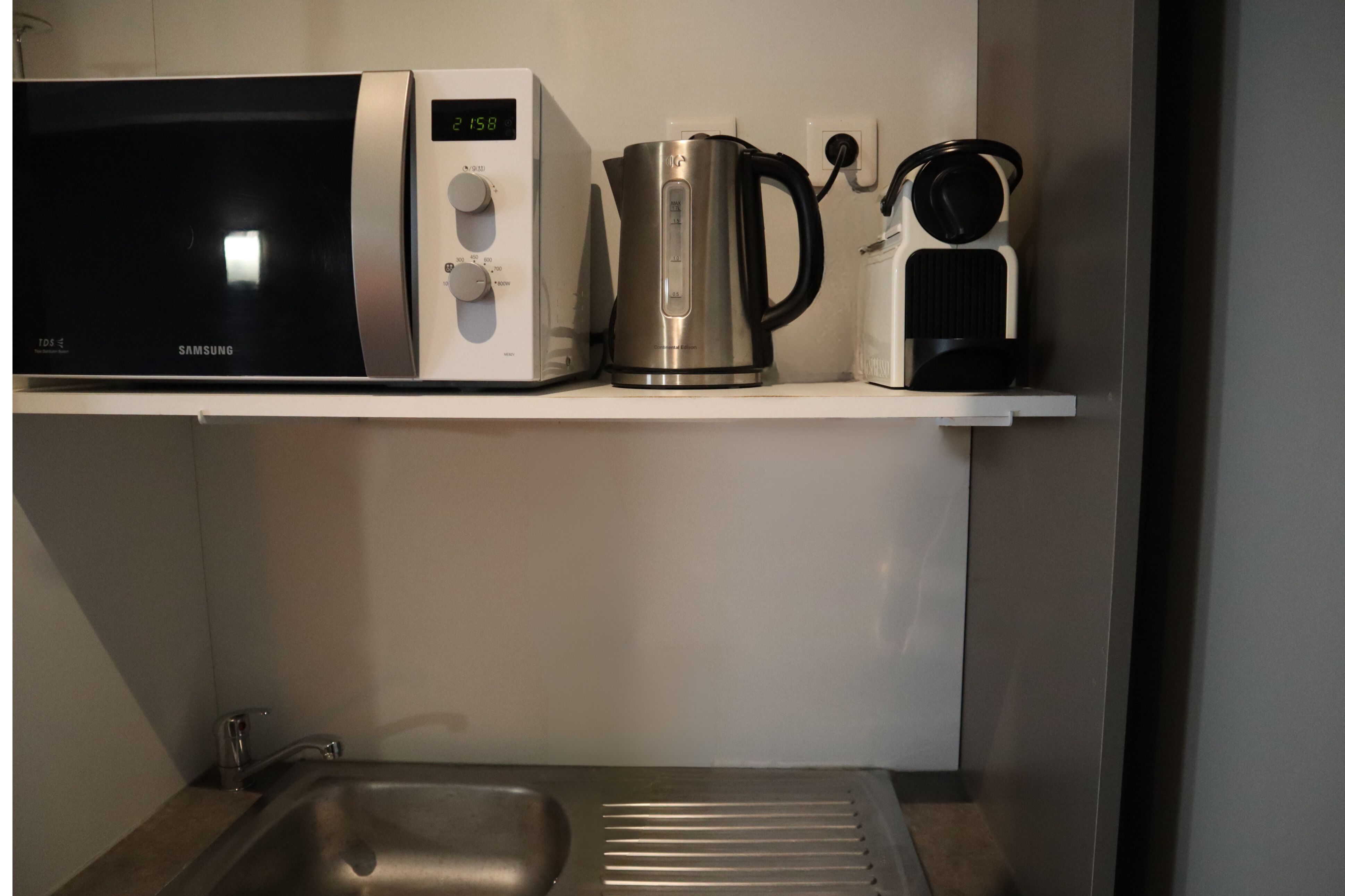 Une cuisine équipée d’un four à micro-ondes, d’une machine à café et d’une bouilloire, le tout posé sur une étagère blanche.
