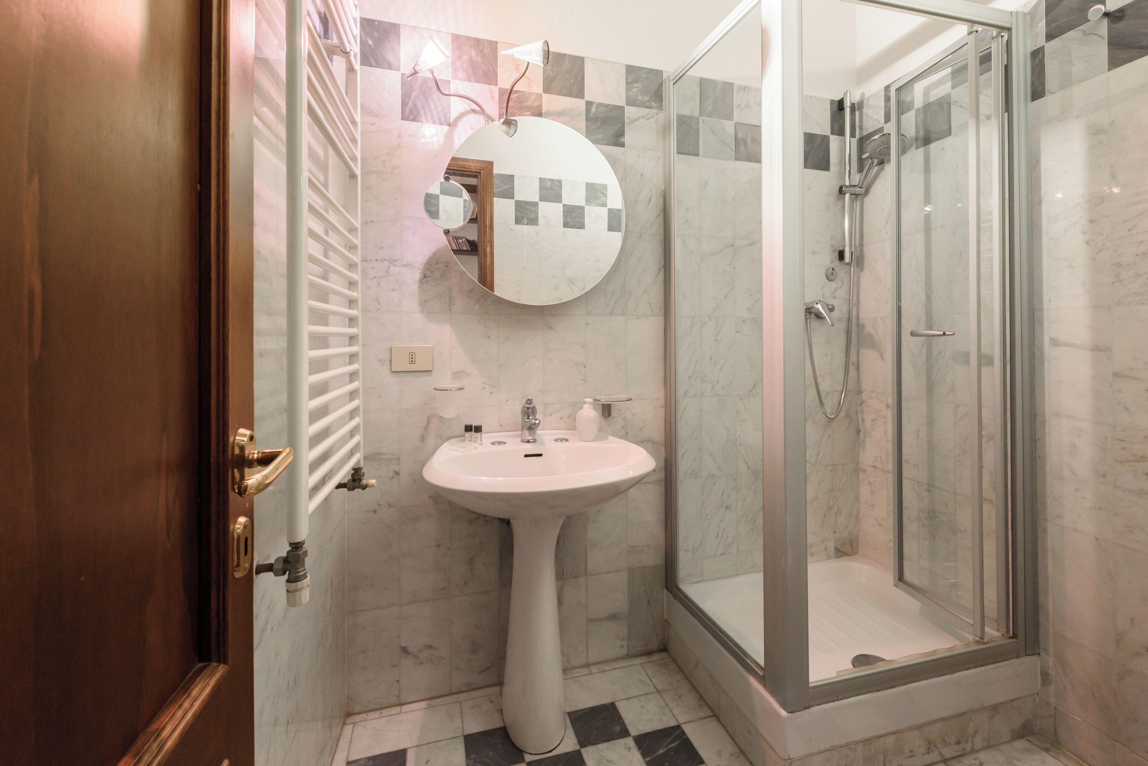 A bathroom with a pedestal sink, a round mirror, a glass shower enclosure, and a checkered tile backsplash.