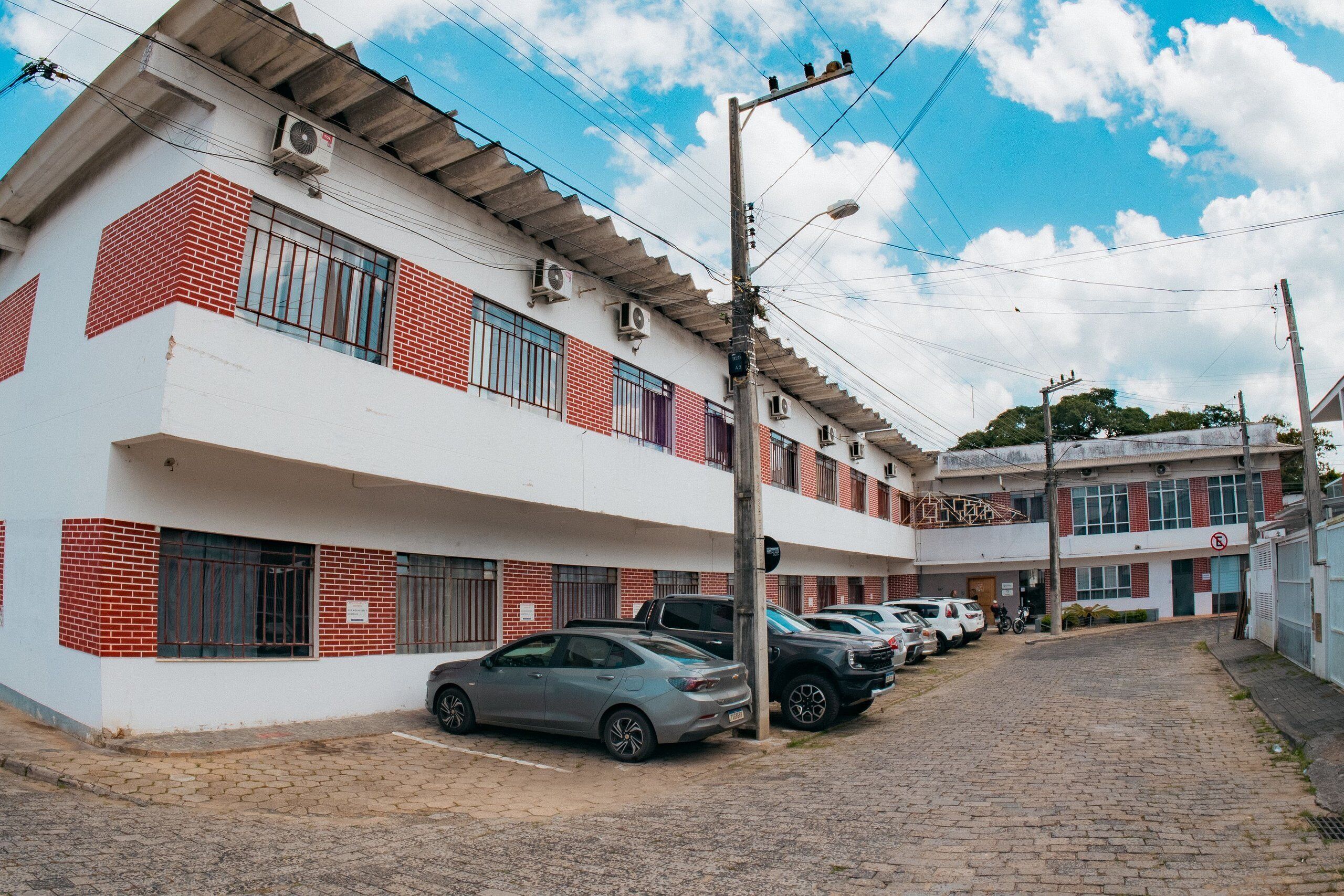 Um edifício de vários andares com fachada branca e detalhes em tijolo vermelho, com várias varandas e uma fileira de carros estacionados.