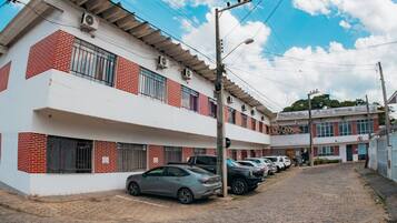 Um edifício de vários andares com fachada branca e detalhes em tijolo vermelho, com várias varandas e uma fileira de carros estacionados.