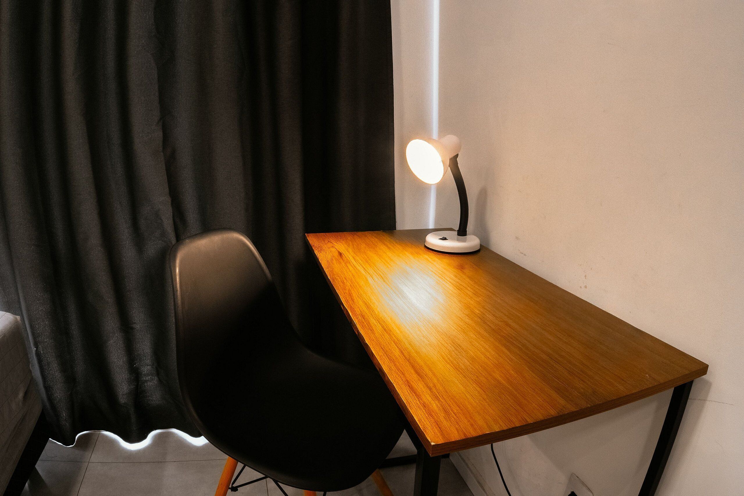 A wooden desk with a lamp, a black chair, and a black curtain in a room.