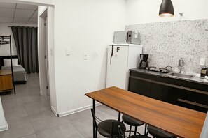 Cozinha compacta com micro-ondas, cafeteira e pia. Há uma mesa de madeira com duas cadeiras. Uma cama beliche é visível no quarto adjacente.