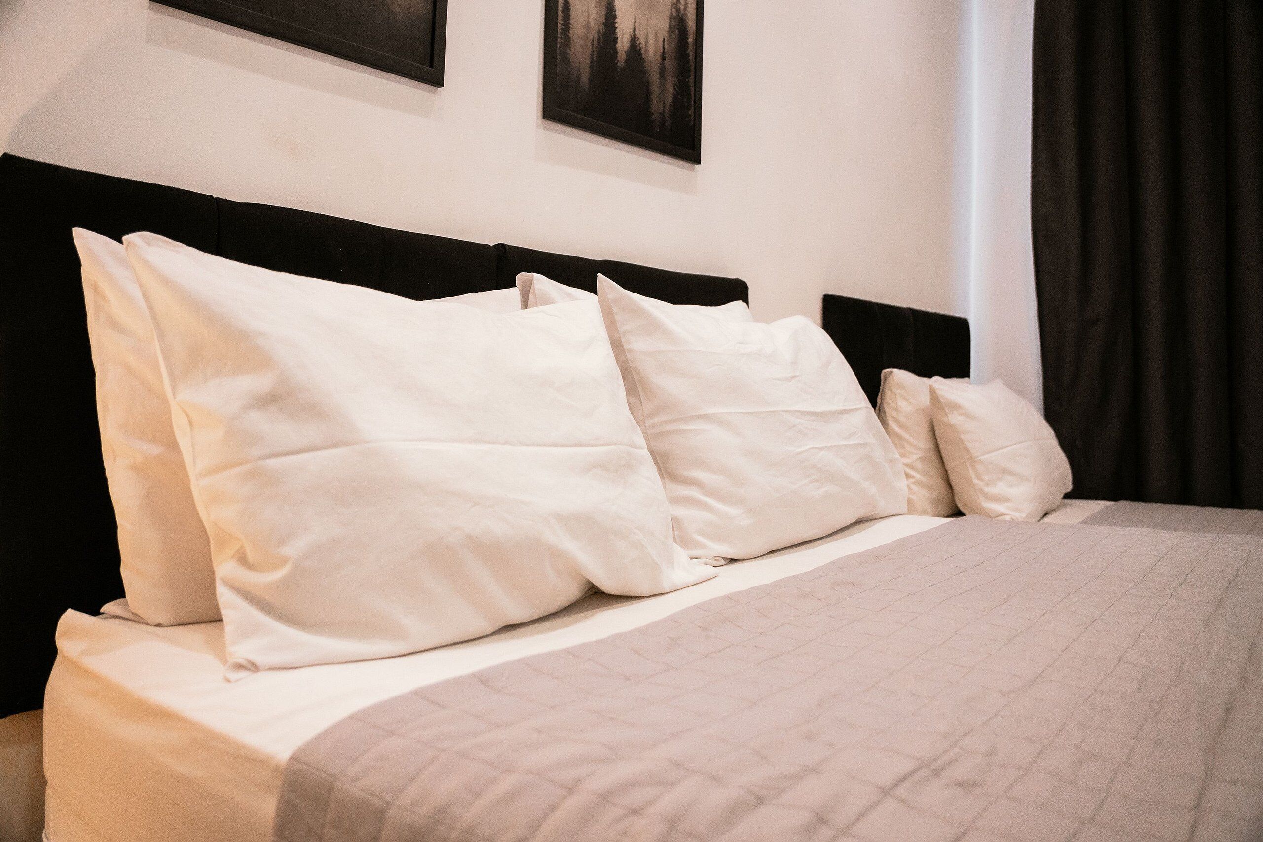 Una cama con cubierta acolchada y varias almohadas, cabecero oscuro y dos cuadros en la pared.