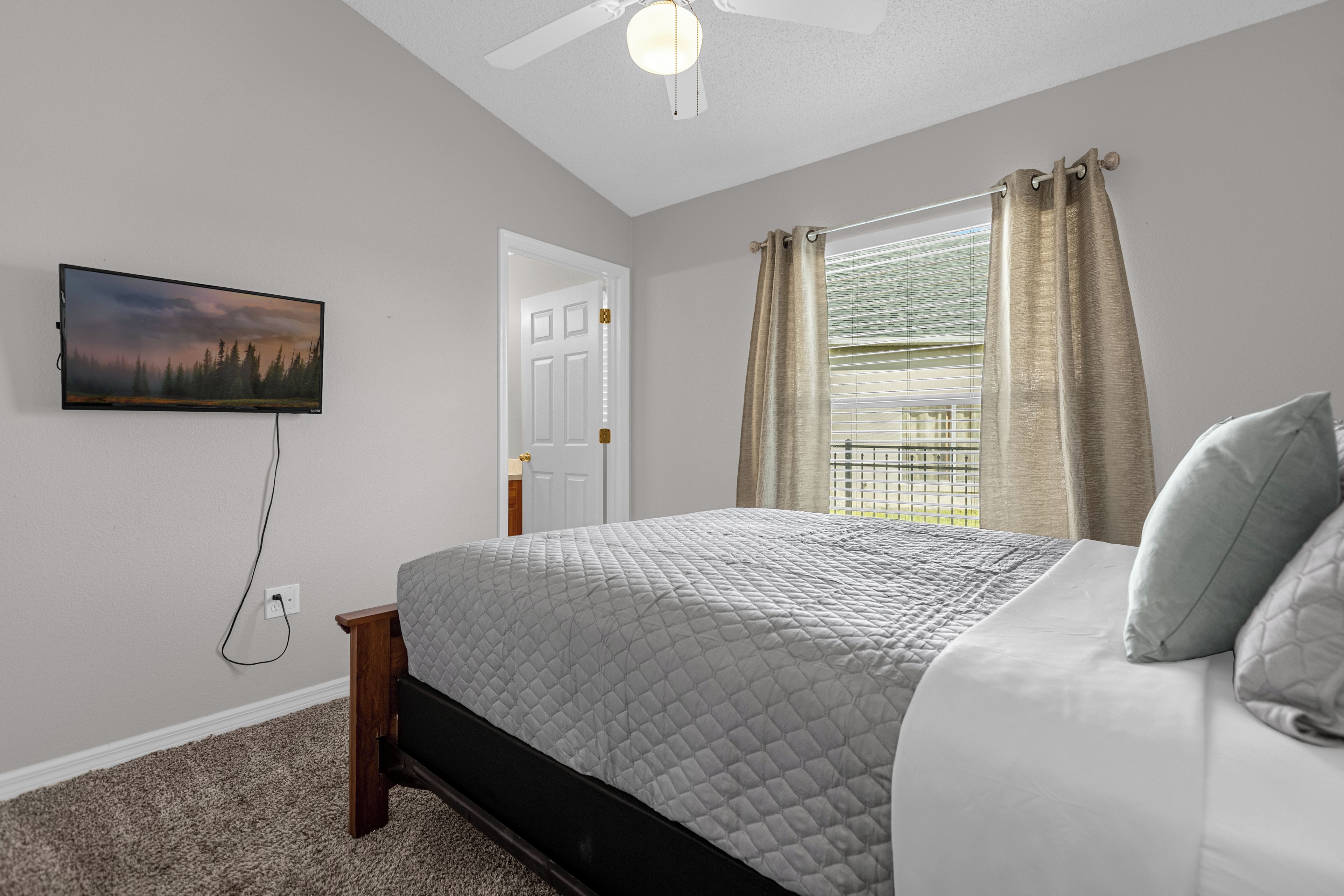 A bedroom with a bed, a television mounted on the wall, a window with curtains, and a ceiling fan.