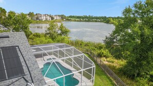 Una casa con piscina, rodeada por una valla blanca, con vista a un lago con árboles y edificios al fondo.