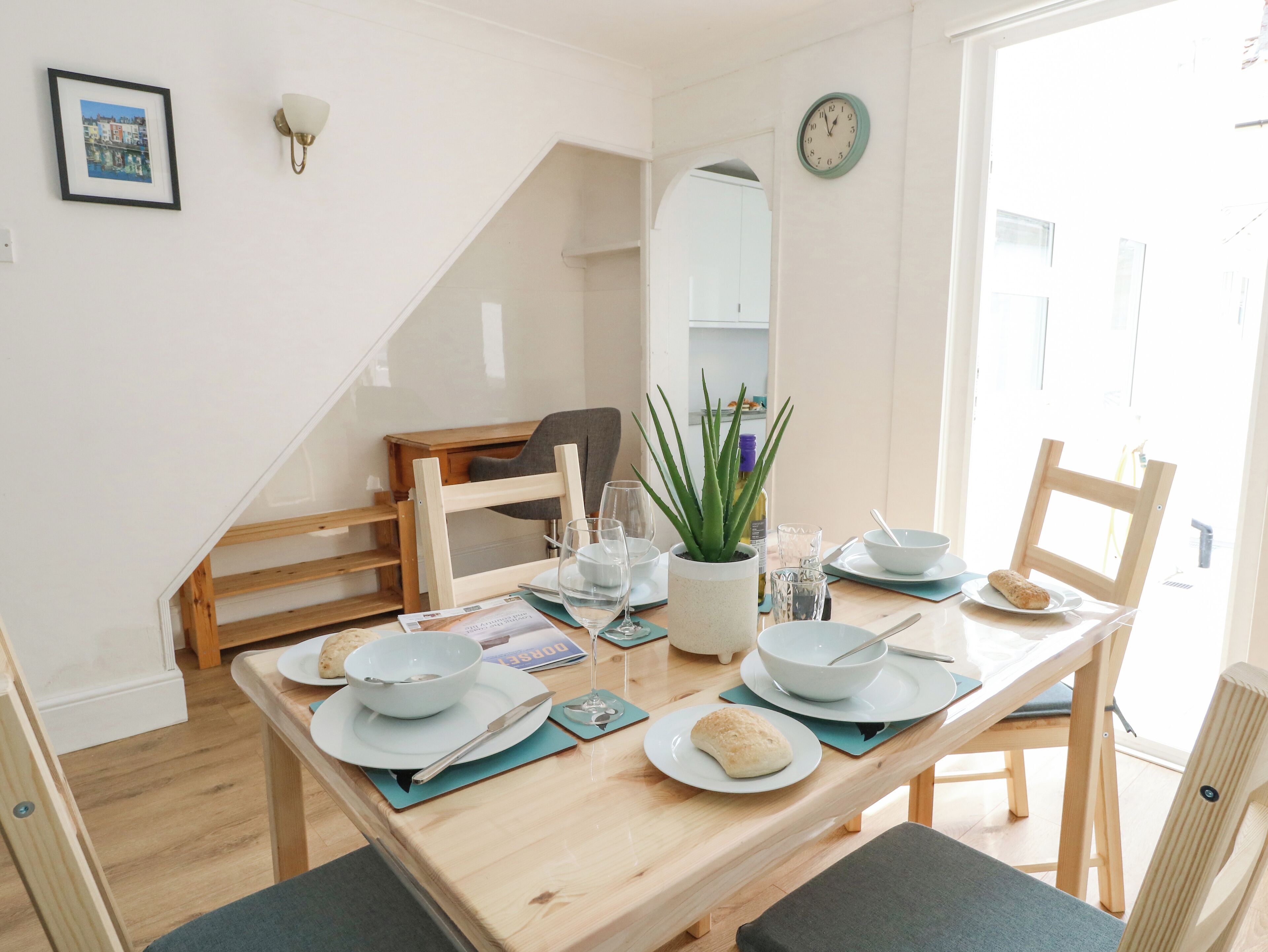 A dining area with a wooden table set for two, a potted plant, and a clock on the wall.