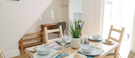 A dining area with a wooden table set for two, a potted plant, and a clock on the wall.