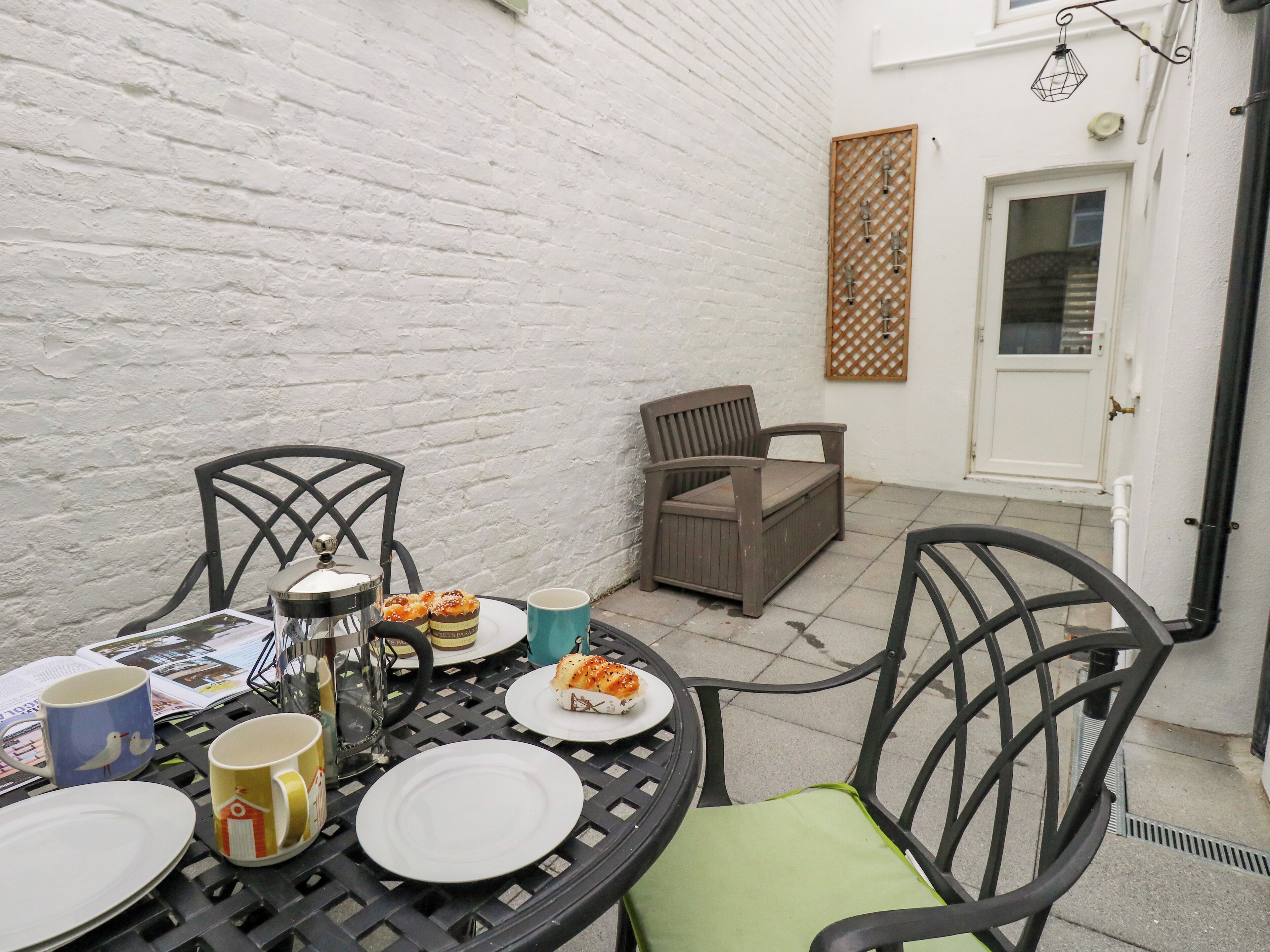 A patio with a round table set for two, two chairs, a small table with a teapot and cups, and a door with a lattice panel.