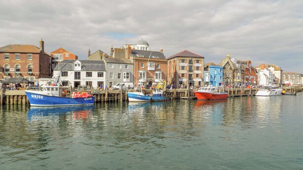 A waterfront with several boats docked, a row of buildings with various businesses, and a clear sky. - Day Cottage (Weymouth)