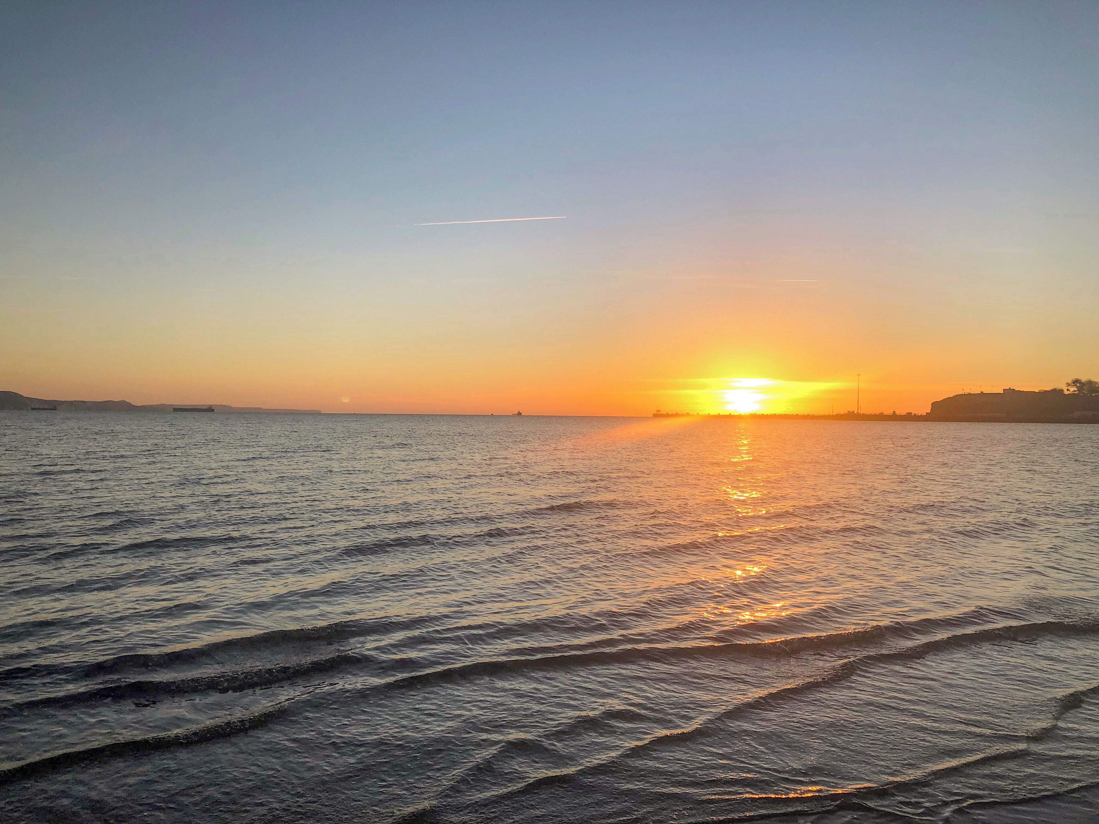 Un coucher de soleil sur une mer calme aux vagues douces, avec une côte lointaine et un ciel dégagé.