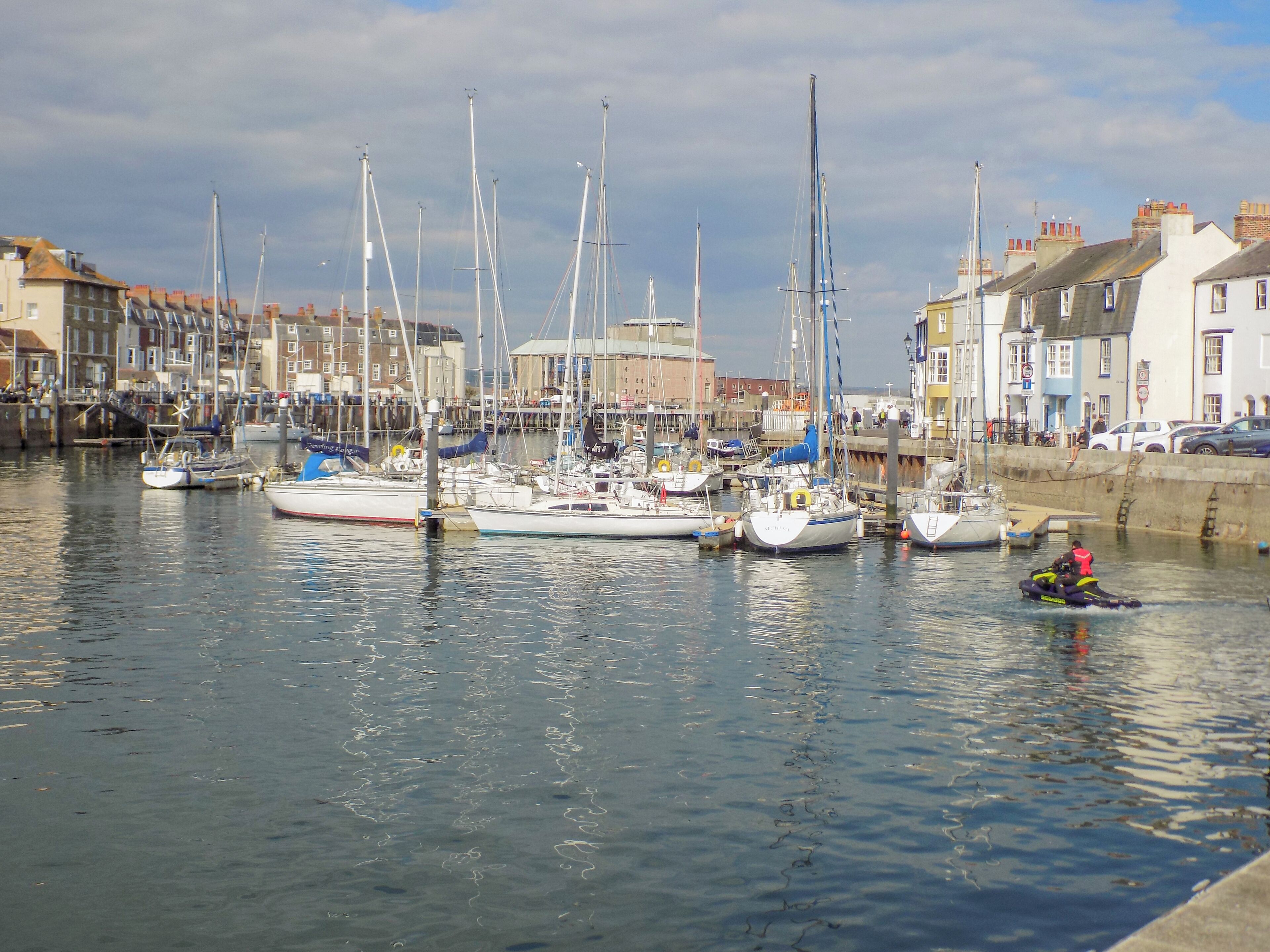 Un port de plaisance où sont amarrés différents types de bateaux, un jet ski sur l’eau et des bâtiments le long de la rive.