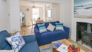 A living room with a blue sofa, a wooden coffee table with a magazine and fruit, and a painting of a beach scene. - Day Cottage (Weymouth)