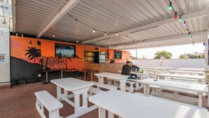 Un espace extérieur aménagé en terrasse avec des tables et des bancs blancs, un coin bar doté d’un écran de télévision et une fresque murale.