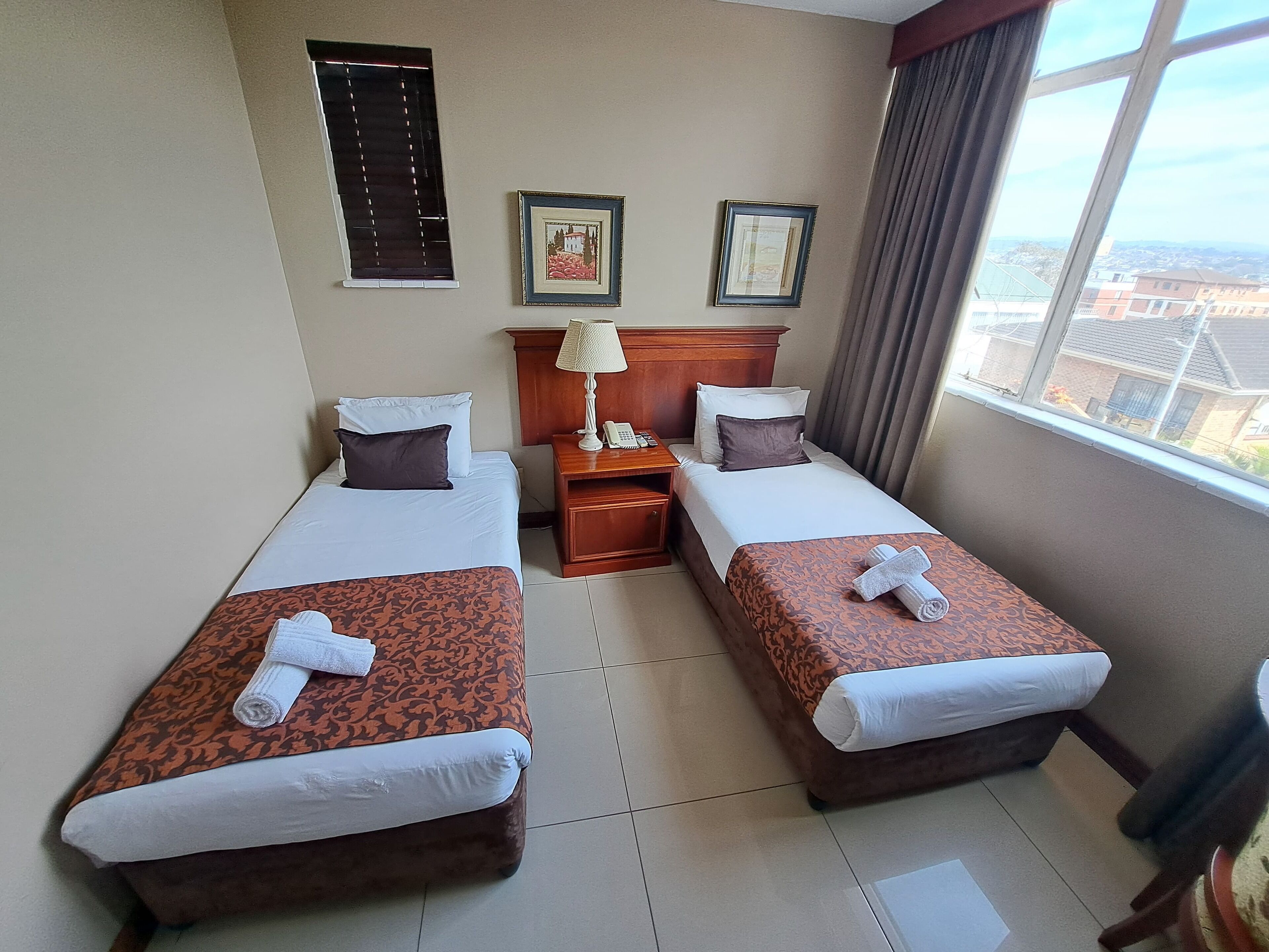 A hotel room with two beds, a nightstand, a lamp, and a window with a city view.