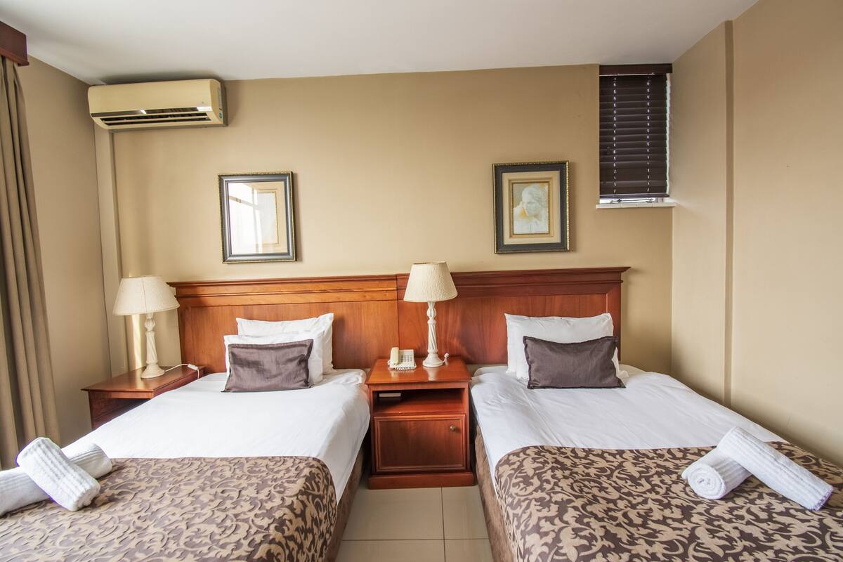 Une chambre d’hôtel avec deux lits, une tête de lit en bois, des lampes de chevet, un téléphone et une fenêtre avec des stores.