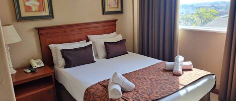 A hotel room with a large bed, a nightstand with a phone and remote controls, two framed pictures on the wall, and a view of the outside through a window with curtains.