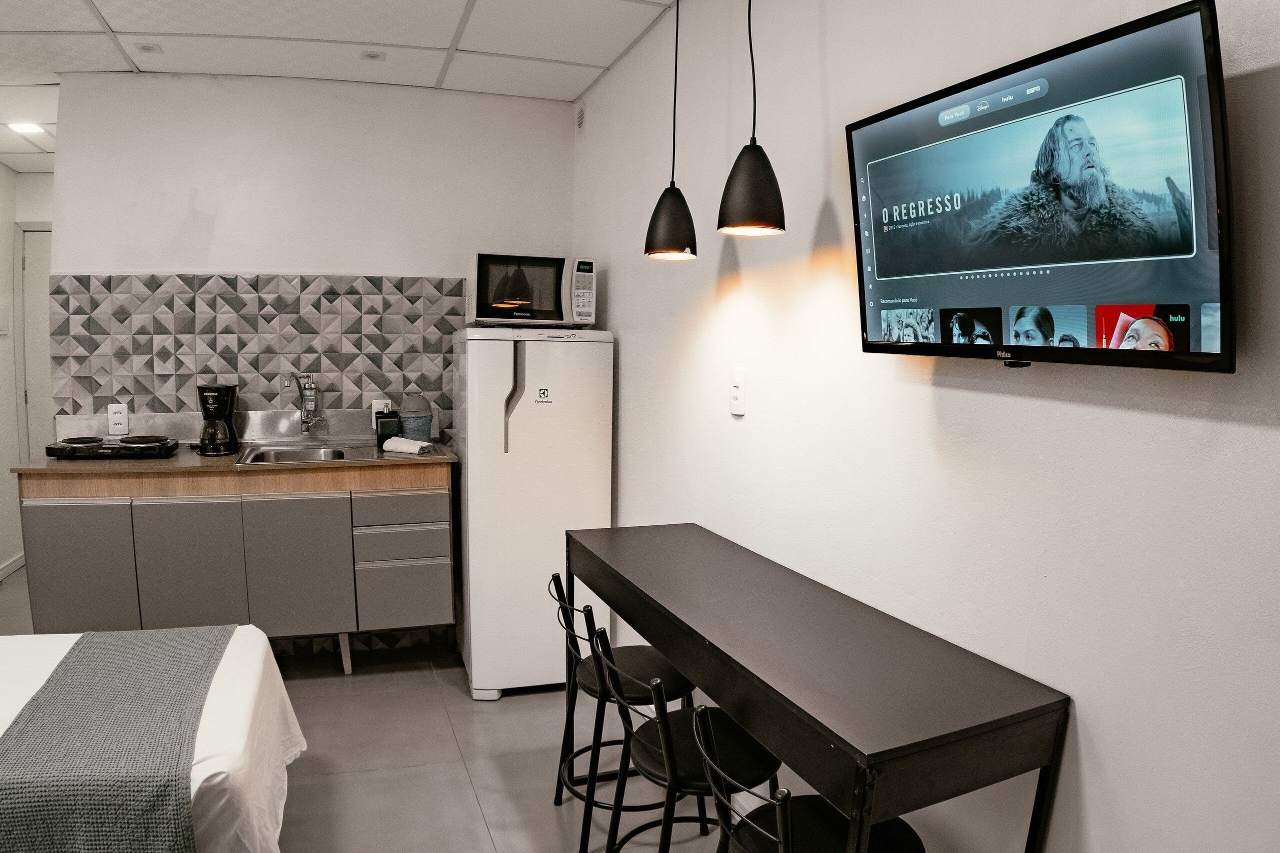 A modern kitchen with a microwave, coffee maker, and refrigerator. There is a dining table with chairs and a TV mounted on the wall displaying a movie.