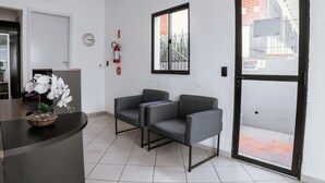 Un área de recepción con un mostrador, dos sillones grises, un reloj, un extintor y una ventana con vistas a un edificio.