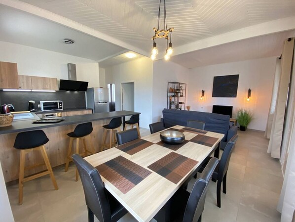 A modern kitchen with a central island, a dining area with a large table and chairs, and a living area with a sofa and a TV.