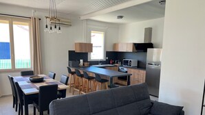 Een moderne keuken met een centraal eiland, houten kasten en roestvrijstalen apparatuur. Er is een zithoek met een tafel en stoelen, en een grijze bank op de voorgrond.