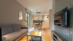 Una sala de estar moderna con sofá, mesa de centro y televisor. Hay un comedor con sillas y una mesa. La habitación tiene ventanas grandes y una cocina al fondo.