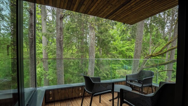 Terraza de madera con barandilla de vidrio que da a un bosque. Hay dos sillas negras y una mesita.