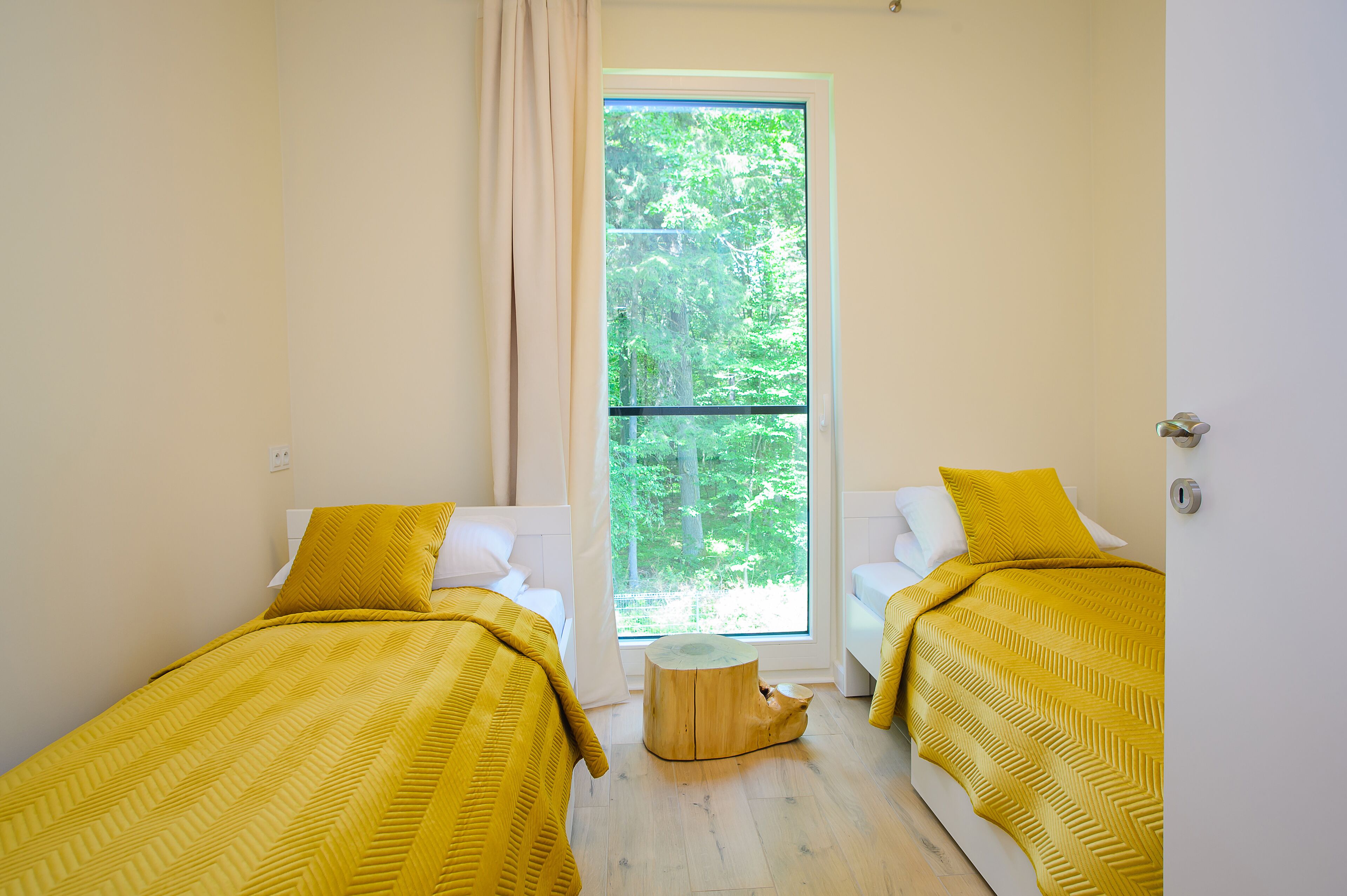 Ein Zimmer mit zwei Betten, jeweils mit einer gelben Tagesdecke, einem runden Holzschemel und einem großen Fenster mit Blick auf Bäume.