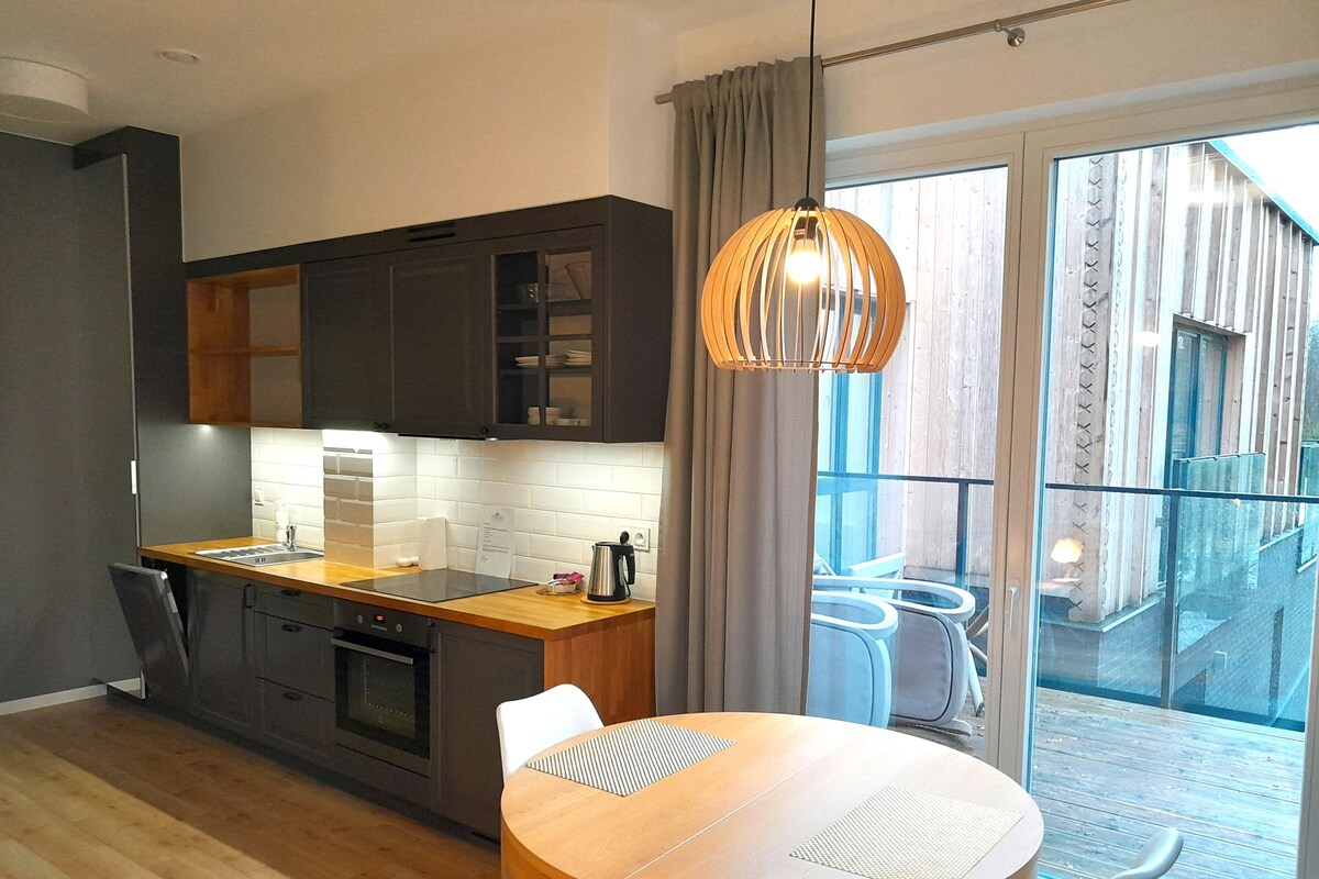 Una cocina moderna con gabinetes oscuros, una encimera de madera y un horno empotrado. Hay una zona de comedor con una mesa redonda y sillas, además de un ventanal con cortinas.