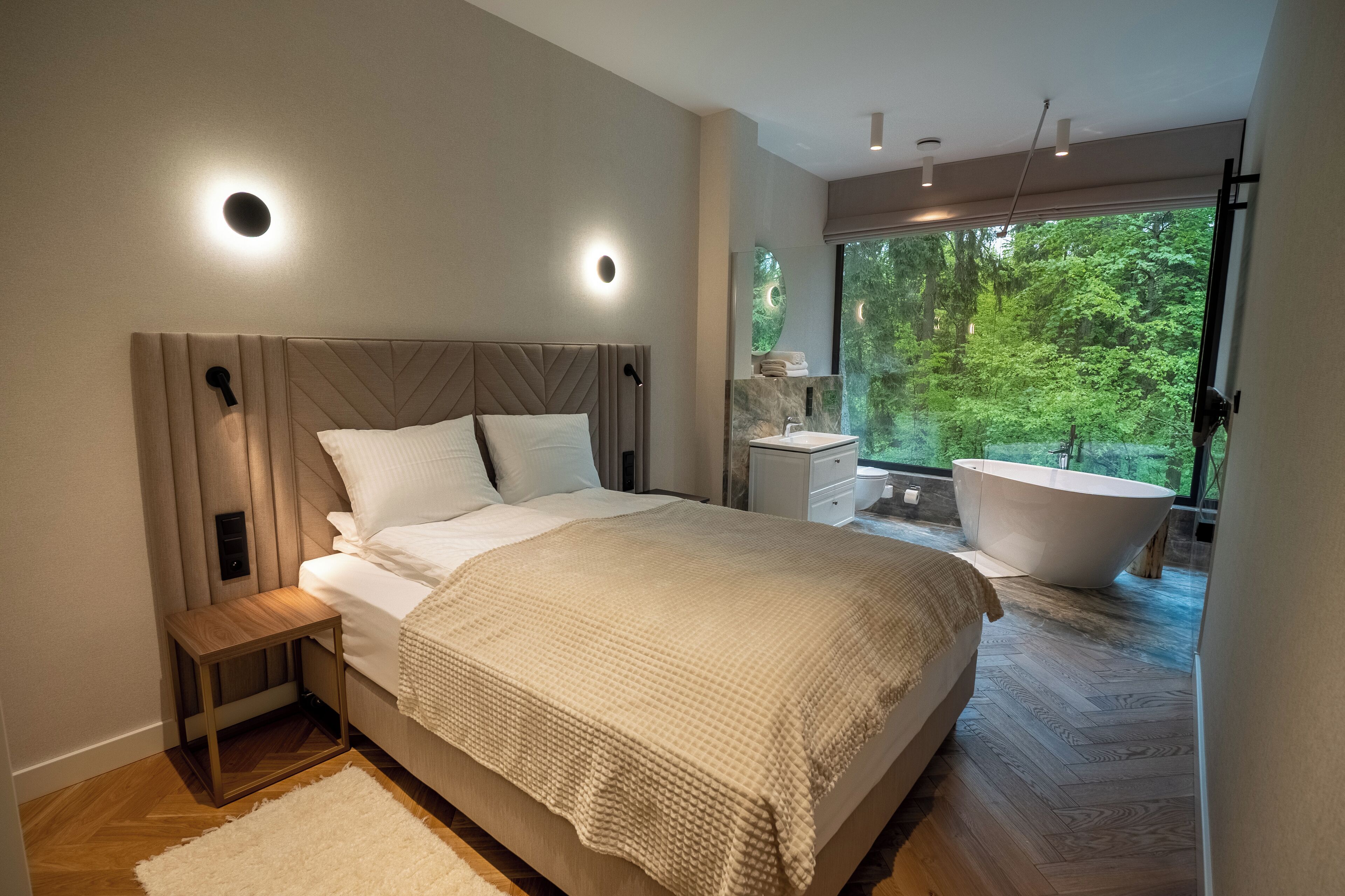 A modern bedroom with a large bed, bedside tables, a built-in bathtub, and a view of lush greenery.