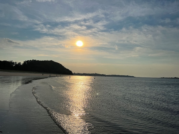 Una playa con un mar tranquilo que refleja el sol, una orilla arenosa y una colina distante con árboles.