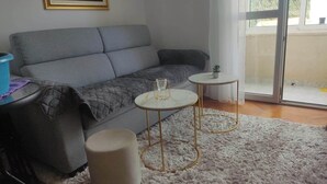 Ett vardagsrum med en grå soffa, två små, runda bord, en vit matta och en skjutdörr i glas som leder ut till ett utomhusområde.