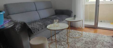 A living room with a grey sofa, two small round tables, a white rug, and a sliding glass door leading to an outdoor area.