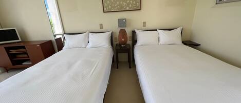A hotel room with two single beds, a desk with a computer, a small table with a lamp, and a wall-mounted picture.