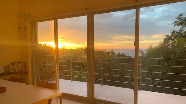 Een kamer met een grote schuifdeur die leidt naar een balkon met uitzicht op de ondergaande zon boven een landschap met bomen en water.