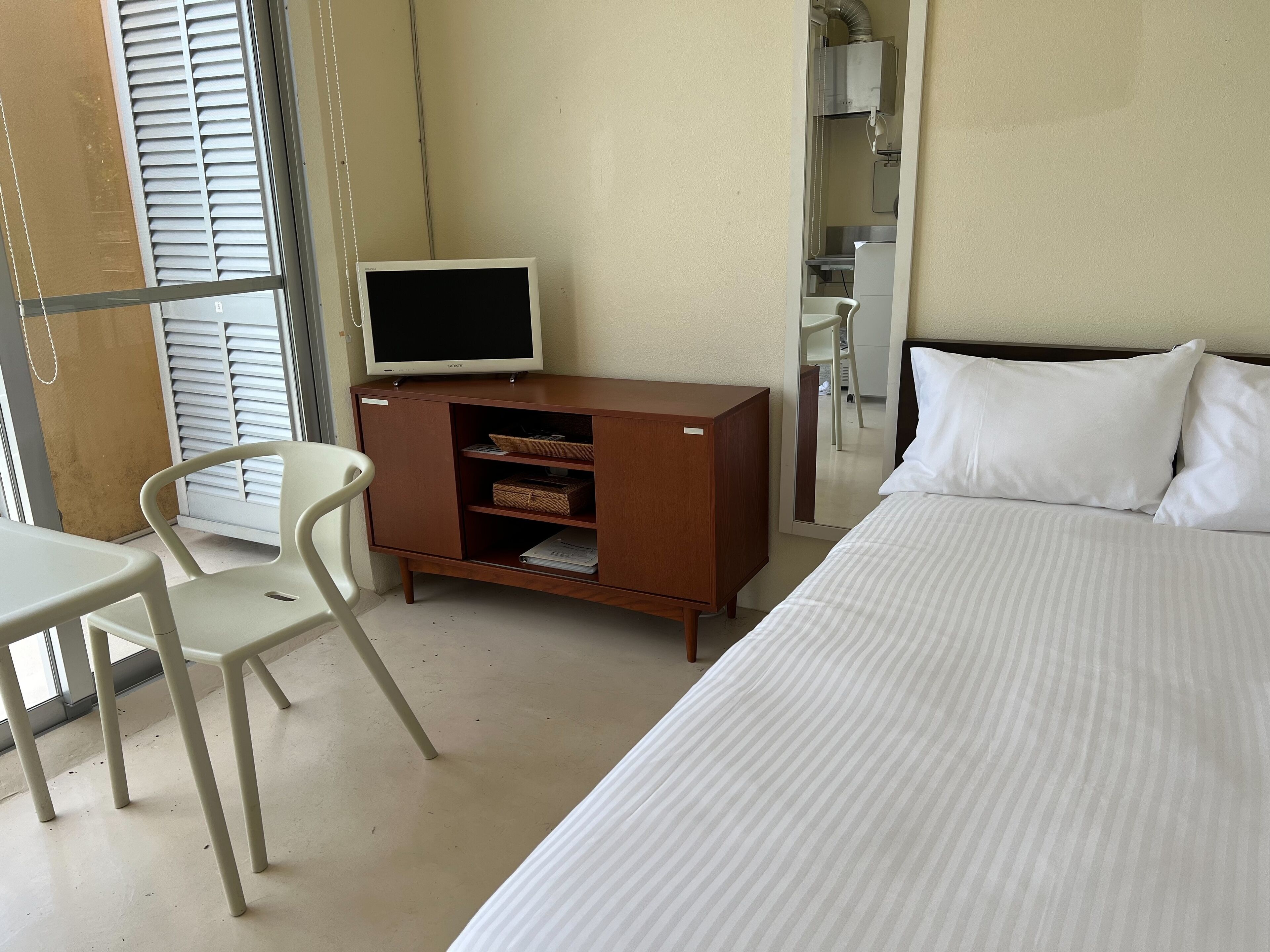 A hotel room with a bed, a TV on a wooden cabinet, a white chair, and a small table.