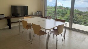 Een ruime kamer met een grote tafel en stoelen, een televisie en uitzicht op de oceaan door grote ramen.