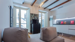 A modern living room with a sofa, a flat-screen TV, and a kitchen area with white cabinets and a countertop. - Casa Vel Apartment by Wonderful Italy (Turin)