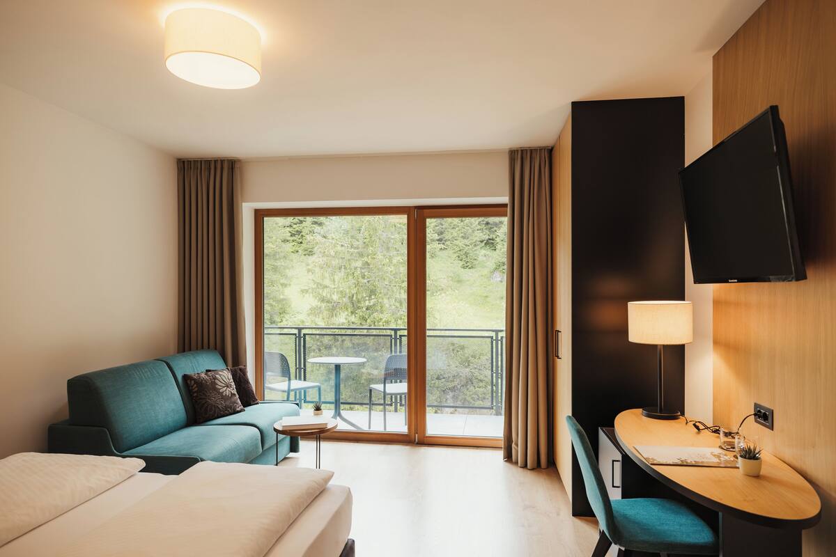 Hotellihuone, jossa on sänky, työpöytä tuolilla, sohva, televisio ja parveke, jolta on näkymä vehreään maisemaan.