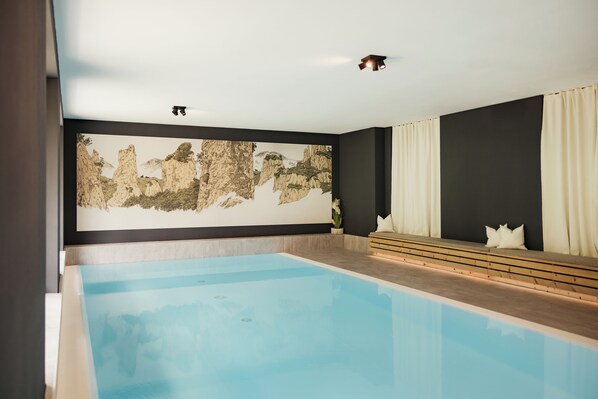 Una piscina interior moderna con un diseño minimalista, que incluye un gran mural en la pared, una zona de estar con cojines y un espacio limpio y despejado.