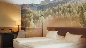 Habitación de hotel con cama, escritorio y lámpara. Amplia ventana con vistas a un bosque y montañas.