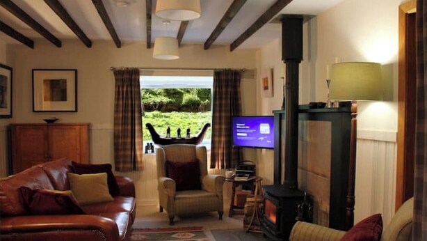 A cozy living room with a fireplace, leather sofa, armchair, and a television. - Smithy Cottage Licence AS00607 (Strathdon)