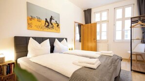 Cama doble con sábanas blancas, cabecera gris, un cuadro sobre la cama, una puerta de madera y una ventana con cortinas.