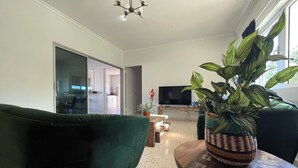 Ein Wohnzimmer mit einem grünen Sofa, einem Fernseher auf einem Schrank, einer Pflanze auf einem Tisch und einer Glastür, die in eine Küche führt.