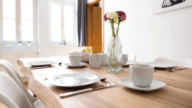 A dining table set with plates, cups, and cutlery, a vase with flowers, and a window with curtains. - Ko-Living Space Opera | Suite 6 | Kitchen | Living room | Dart | City center (Halle (Saale))