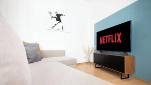 A modern living room with a large flat-screen TV mounted on a blue wall, a white sofa, and a wooden TV stand. - Ko-Living Space Opera | Suite 6 | Kitchen | Living room | Dart | City center (Halle (Saale))