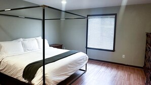 Ein Bett mit Baldachin, ausgestattet mit weißen Leinen und einer dunklen Tagesdecke, in einem Zimmer mit Holzboden und einem Fenster mit Jalousien.