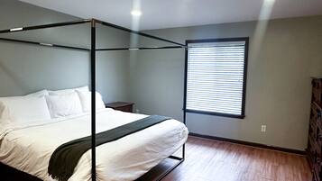 A four-poster bed with white linens and a dark throw blanket in a room with wooden flooring and a window with blinds.